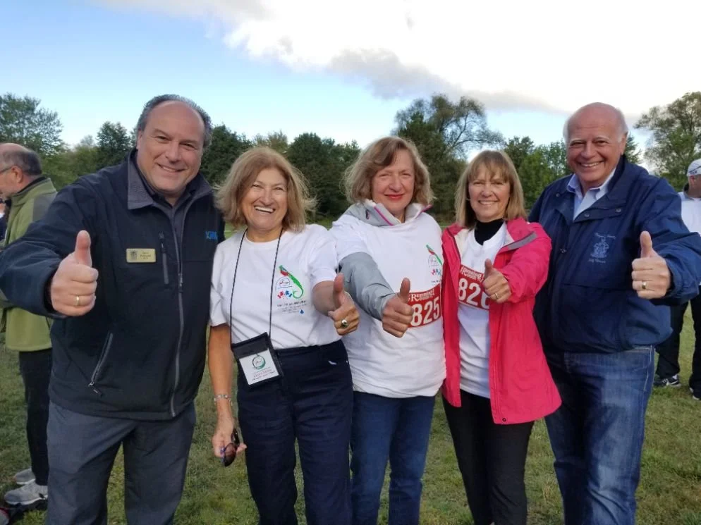 Five smiling people outdoors at a running event, giving thumbs up, some wearing event bibs and casual jackets.
