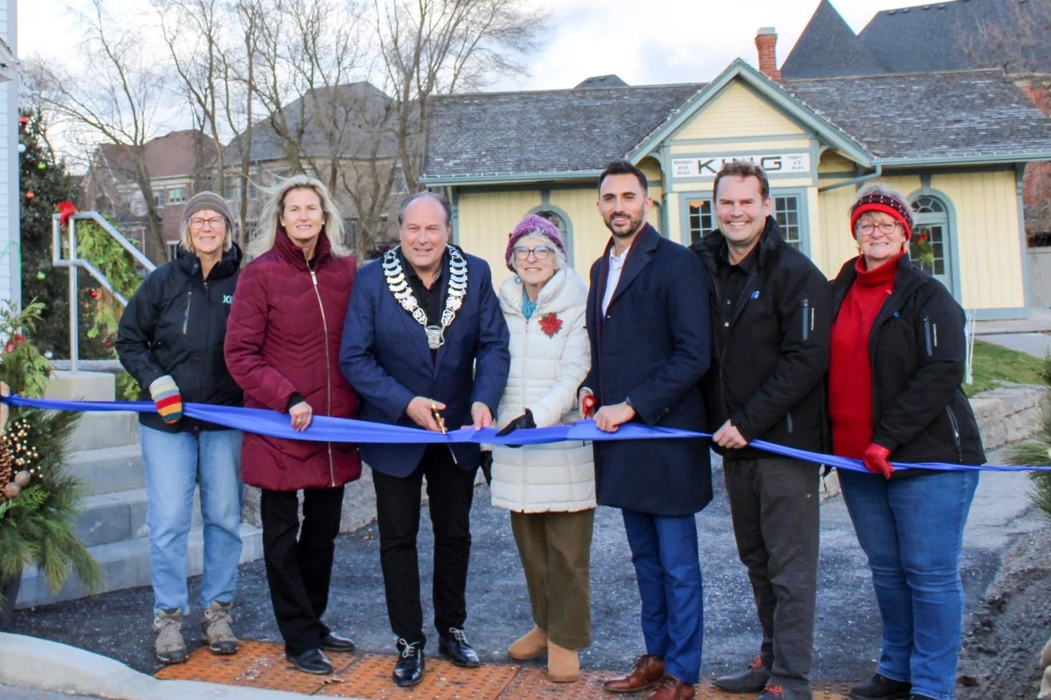 A group of seven people at a ribbon-cutting ceremony outside a small building decorated with Christmas ornaments. The people are smiling and wearing winter clothing. One person wears a mayor's chain, indicating an official role.