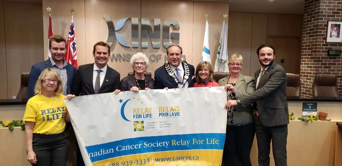 Group of nine people standing in a council chamber, holding a banner that reads "Relay for Life" and "Canadian Cancer Society." They are smiling and dressed in business and casual attire, with flags in the background and the council name visible on t