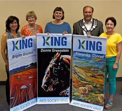 Five women standing indoors, holding arts society banners with their names, smiling.