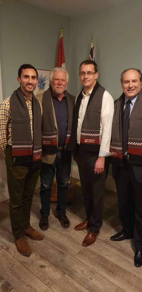 Four men standing together indoors, wearing scarves with a red and black pattern, in front of a wall with flags and artwork.