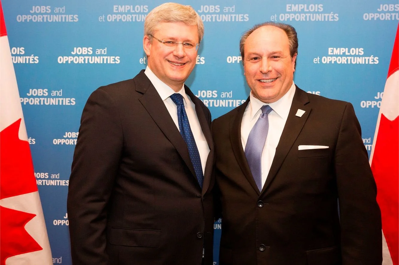 Two men in suits smiling at the camera at a political event with a blue backdrop displaying the text 'Jobs and Opportunities' in both English and French, and Canadian flags on either side.
