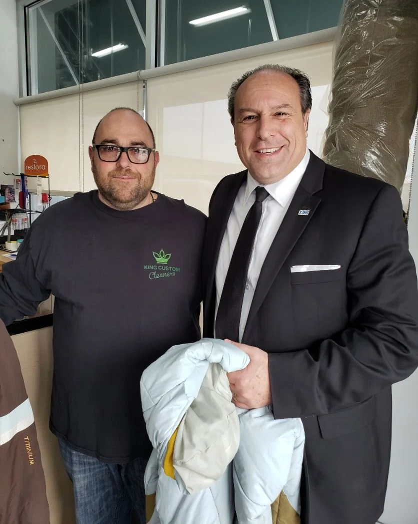 Two men standing indoors, one in a black suit and the other in a black t-shirt with a logo, smiling and holding a white fabric or garment. The background shows windows and office or commercial space decor.
