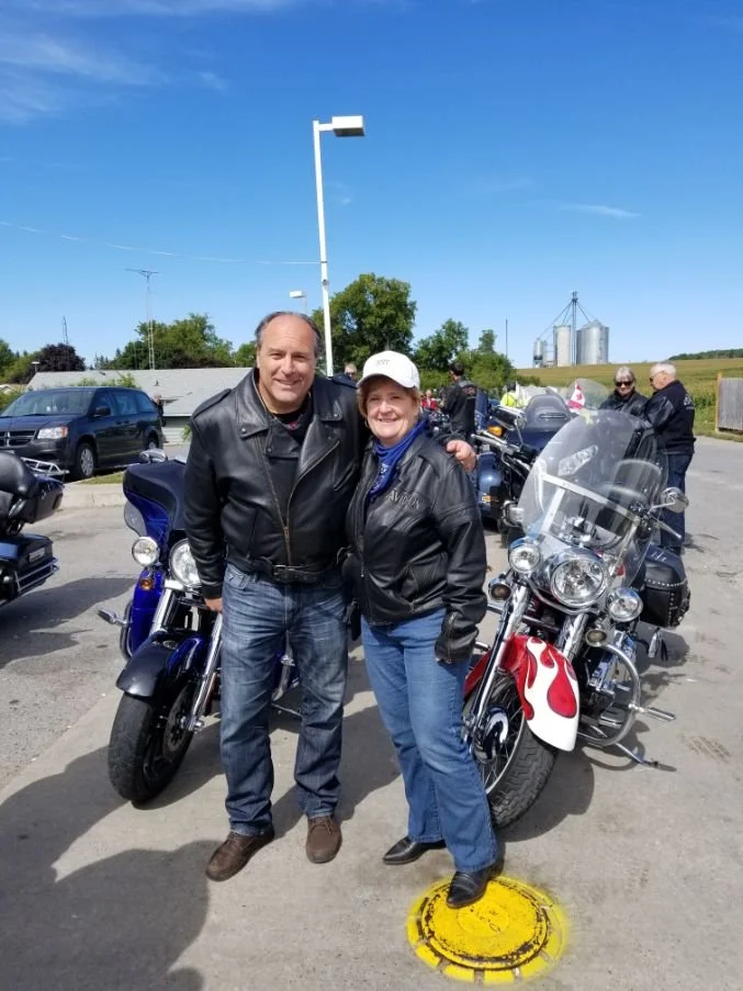 A man and woman wearing leather jackets posing with motorcycles outdoors on a sunny day, with other motorcyclists and rural scenery in the background.