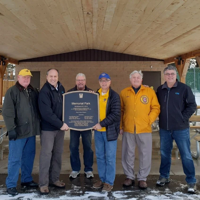 	
Mayor Steve Pellegrini standing outdoors under a wooden shelter holding a memorial park plaque. Winter setting with snow on the ground.