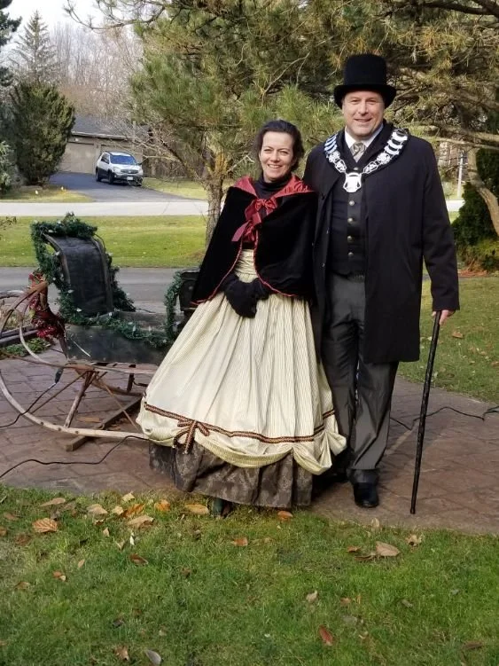 	
Mayor Steve Pellegrini and wife dressed in historical clothing standing outdoors in front of a decorated sleigh, with trees and houses in the background.