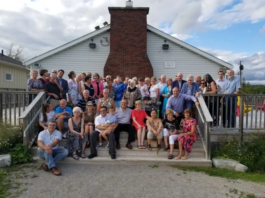 A large group of people gathered on the steps in front of a building with a brick chimney and white siding, outdoors on a partly cloudy day.