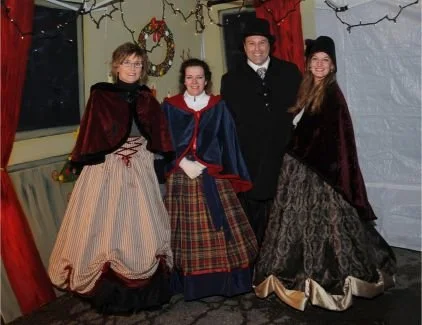 Four people dressed in historical or period costumes standing together in a festive setting with holiday decorations in the background.