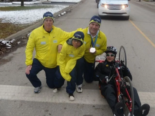 Three men and a person with a specialized bicycle on the side of a road. The men are wearing yellow jackets and beanies with logos, one holding a medal. The person in the wheelchair is wearing sunglasses and a helmet.
