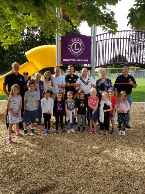 Group of children and adults gathered for a ribbon-cutting ceremony at the Mobileton Lions Club playground with a slide and a sign for the club.