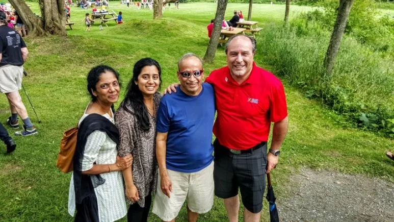 Steve Pellegrini standing outdoors on a grassy area with trees, smiling, with other people in the background.
