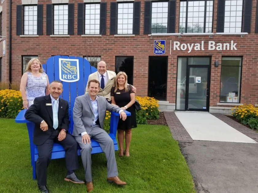 Group of five people posing in front of RBC Royal Bank building, some sitting on a large blue chair, with flowers and grass in the foreground.