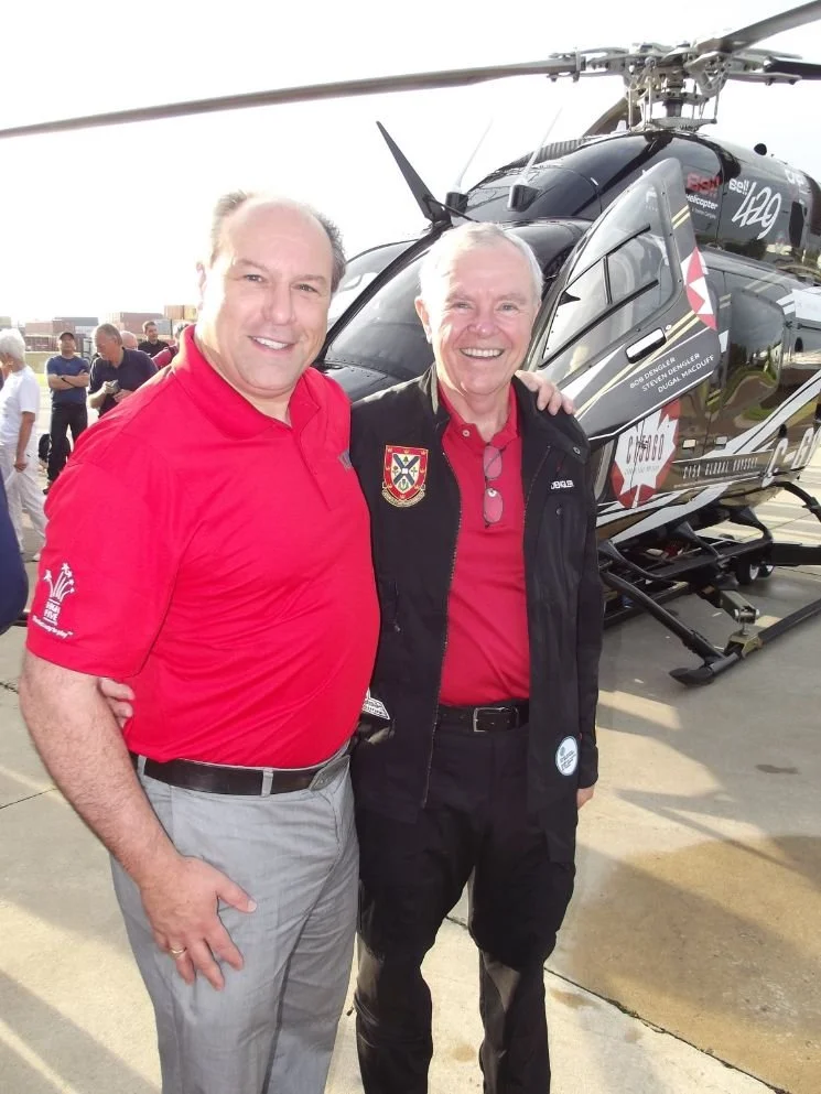 	
Mayor Steve Pellegrini standing in front of a helicopter, smiling at the camera. One man is wearing a red shirt and gray pants, while the other is wearing a black jacket with an emblem and black pants.