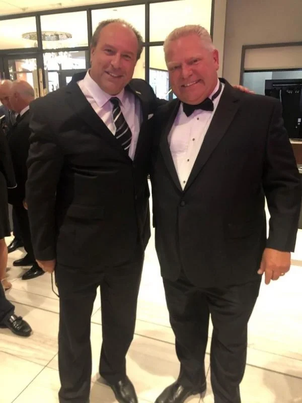 Mayor Steve Pellegrini and Premier Doug Ford dressed in tuxedos standing close together, smiling, with their arms around each other in a formal setting.