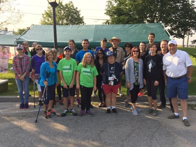 Group of people gathered outdoors for a photo, some wearing matching green shirts that say 'Outrun Cancer,' standing in front of a large green tent, with trees and a lamp post in the background.