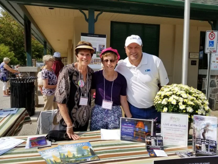 Three people standing behind a table outdoors, smiling at the camera. The table has promotional materials and a large flower arrangement. Other people are visible in the background, and a building with a green door is behind them.