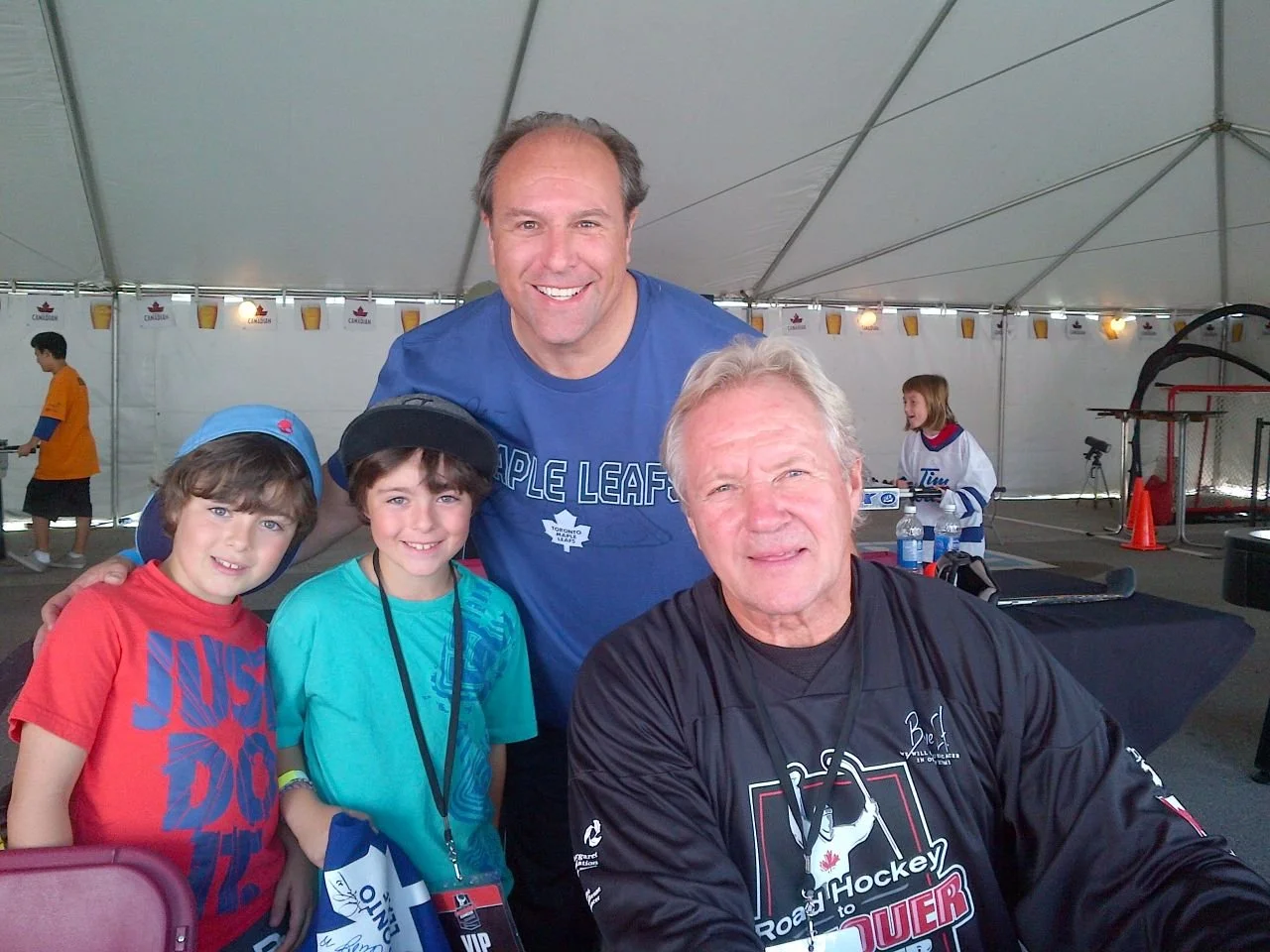Group of four people, two children and two adults, smiling and posing for a photo inside a large tent at an event. The children wear casual clothes and hats, and the adults are wearing hockey jerseys. The background shows other children and event-rel