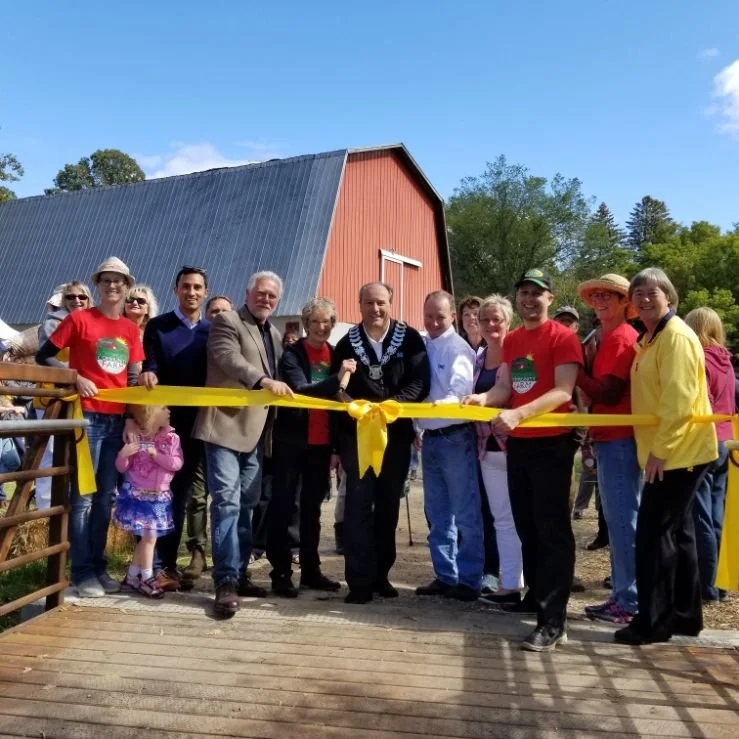 Group of people at a groundbreaking ceremony in front of a barn, holding a yellow ribbon.