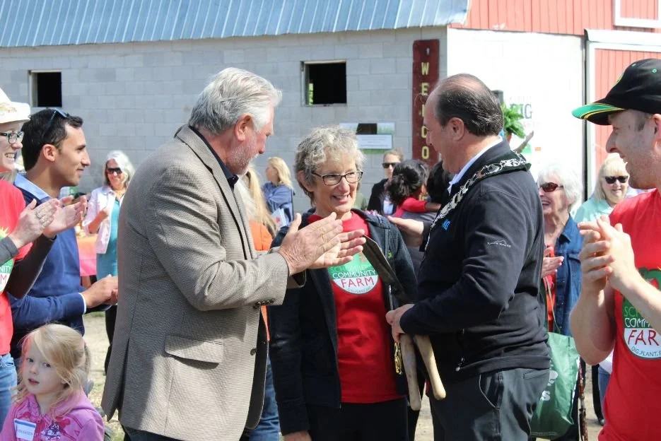 A group of people, including a woman and a man in a sash, at an outdoor farm event, with some clapping and smiling.