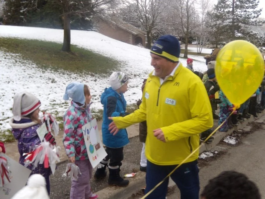 A man in a yellow jacket and navy hat smiling and giving a high five to a young girl holding a sign. Children line up on a sidewalk with snow in the background, some holding signs and balloons.