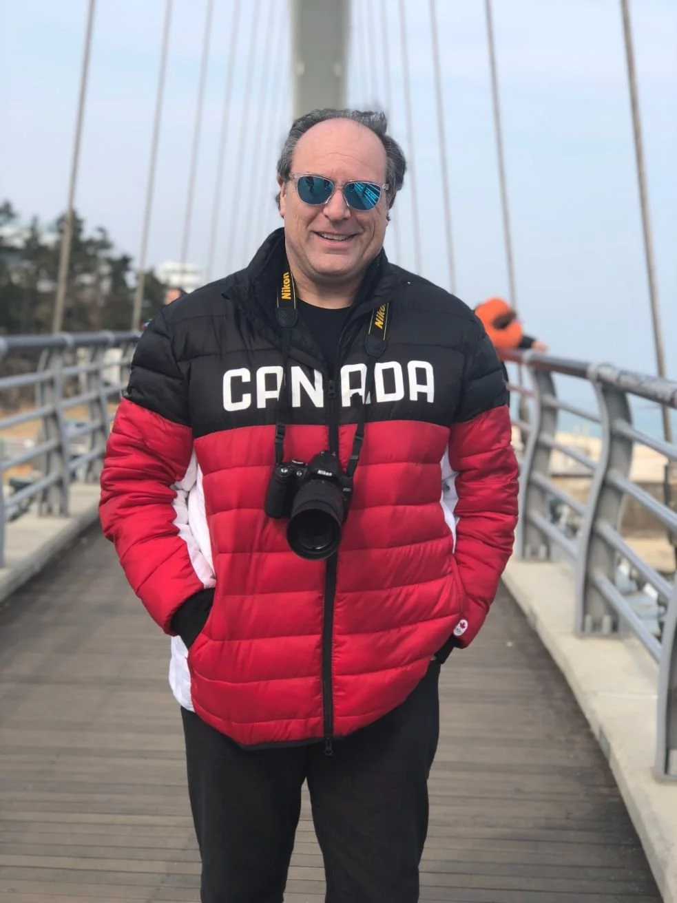 A man standing on a wooden bridge wearing sunglasses, a Canada jacket, and carrying a Nikon camera. The background shows a cloudy sky and some trees.
