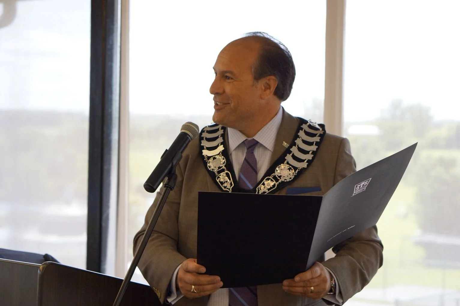 Steve Pellegrini wearing formal attire and a chain of office, standing in front of a microphone, holding a folder, and speaking at an event.
