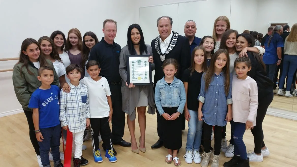 Group of children and adults posing for a photo indoors, with one woman holding a framed award or certificate, some adults wearing formal attire, in a room with a wooden floor and white walls.
