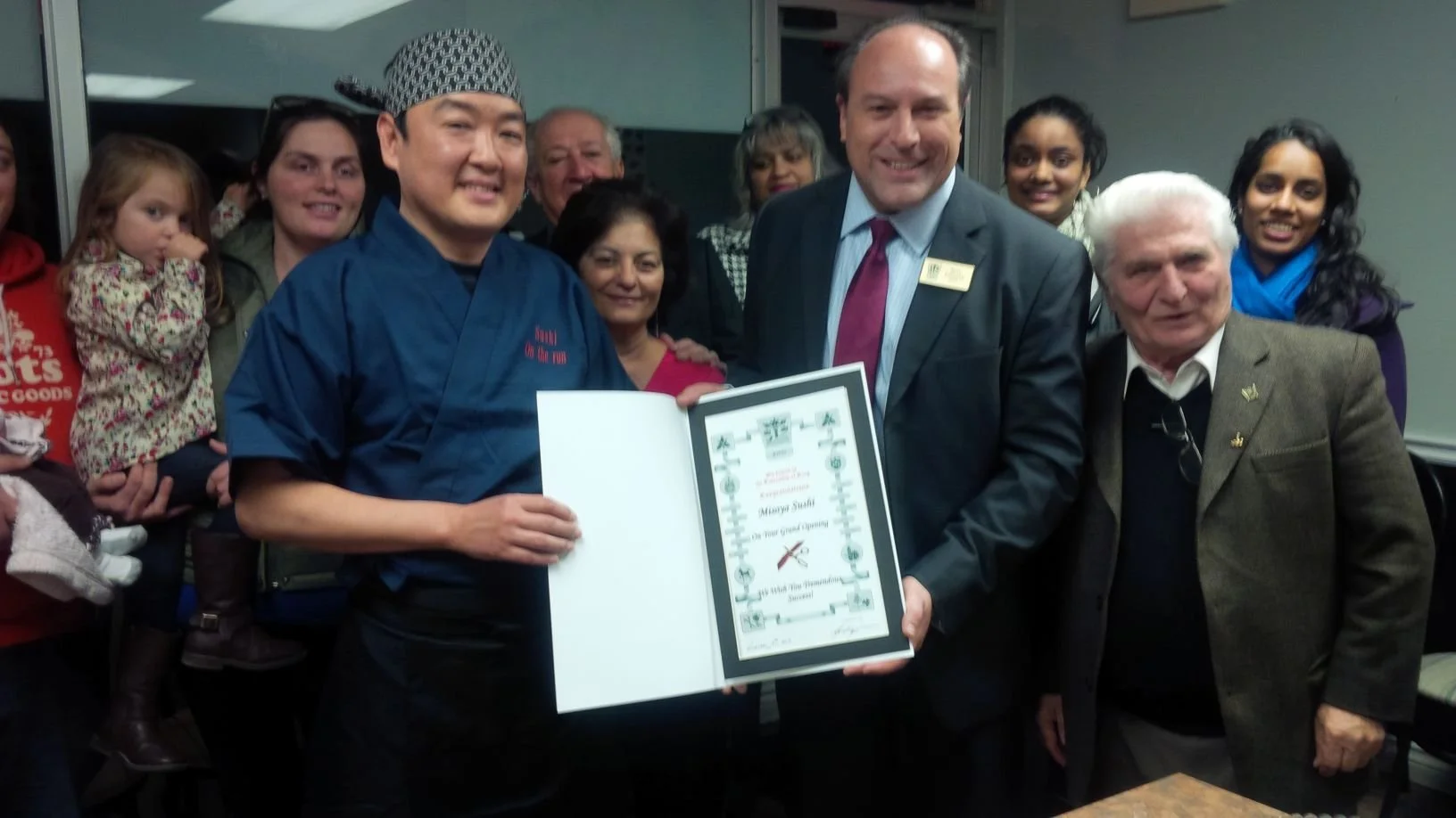Group of people gathered together, with one person holding an award certificate, in a room with window and gray walls.