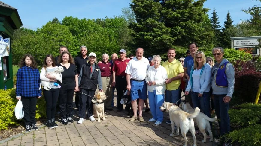 Group of people standing outside on a pathway, holding dogs, with green trees and bushes in the background.