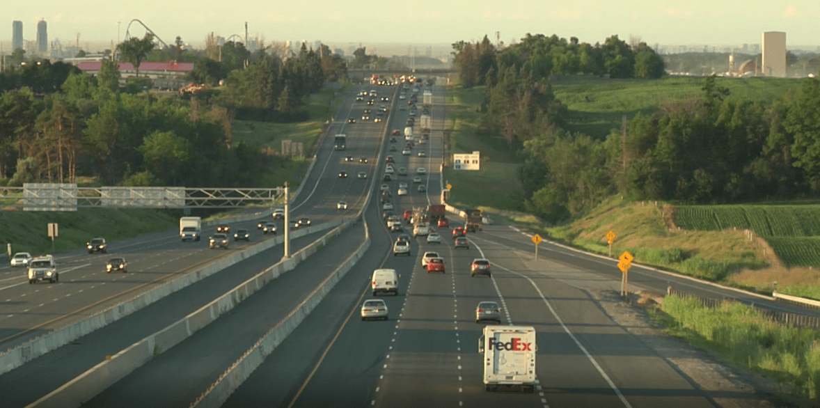 A busy highway with multiple lanes of traffic and various vehicles, including a FedEx truck, during daytime with greenery and office buildings in the background.