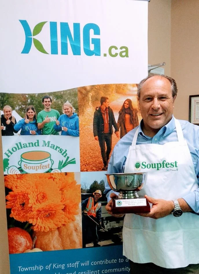 A man wearing a white apron with the 'Soupfest' logo, holding a trophy at an event promoting community and local food, with a 'KING.ca' banner and various photos of people and food in the background.