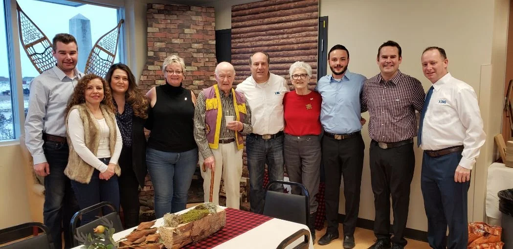 Group of eleven people standing together in a restaurant or banquet room, posing for a photo. There is a table with a decorative centerpiece and wrapped food in front of them.