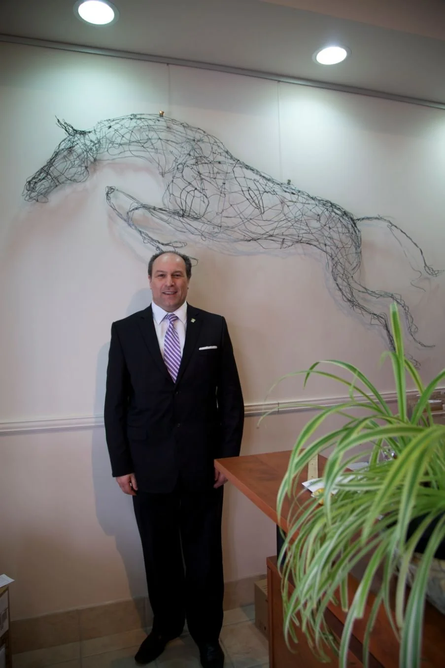 	
Mayor Steve Pellegrini standing in front of a wall with a wire sculpture of a horse's head. There is a wooden table with a green plant partially in the foreground.