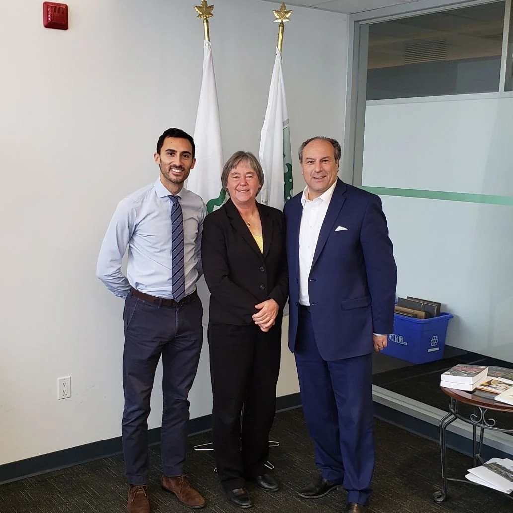 Three professionally dressed people standing together indoors, with two white flags with green symbols behind them, in a modern office or conference room.