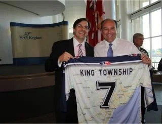 	
Mayor Steve Pellegrini holding a King Township sports jersey with the number 7, smiling at an indoor event with a backdrop and a Canadian flag.