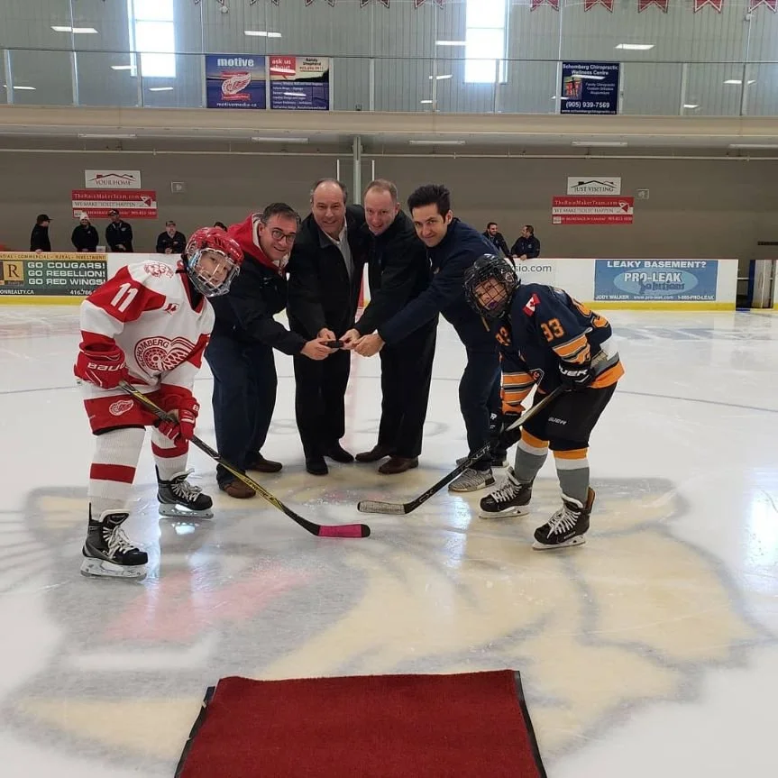 Four people and two hockey players at center ice rink preparing for a ceremonial puck drop.