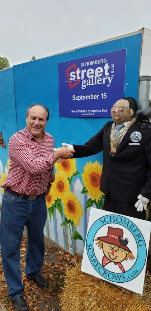 A man in a red checkered shirt and jeans shaking hands with a scarecrow figure dressed in a suit with a gold chain, in front of a colorful sunflower mural at the Schomberg Street Gallery event on September 15, 2019.