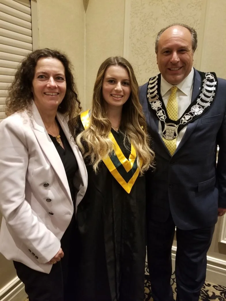 Emma in a graduation gown and sash standing between a Barbara in a white blazer and Mayor Steve Pellegrini in a suit with a chain of office, all smiling for a photo.