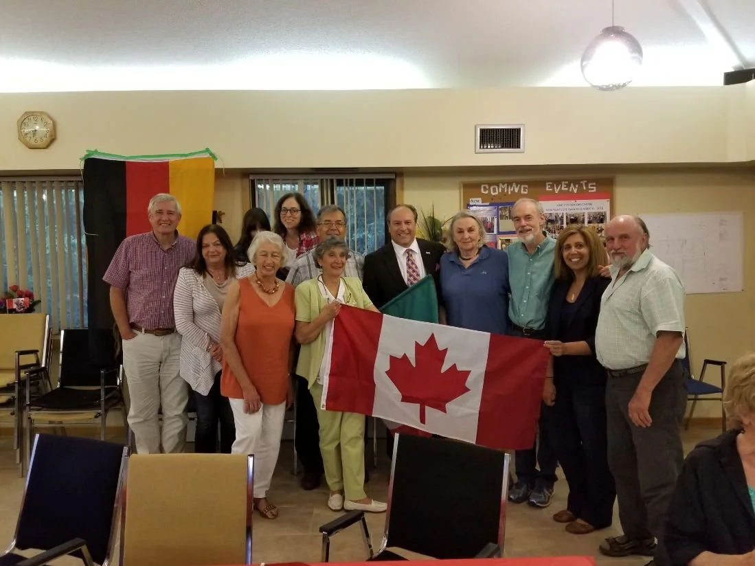 Group of people holding a Canadian flag and standing in a room with a black, red, and yellow flag behind them, celebrating an event.
