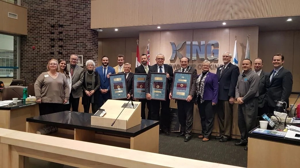 Group of people in a conference room holding framed awards, with a large 'KNG' logo on the wall behind them.