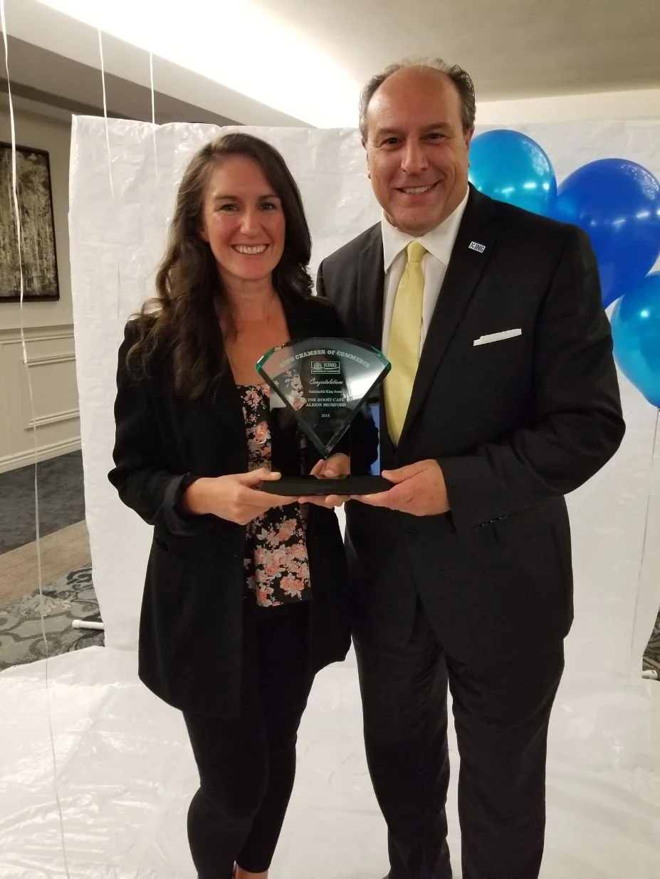 A woman and a man dressed in formal attire, holding a glass award together, smiling at the camera. The background includes a white backdrop and blue balloons.