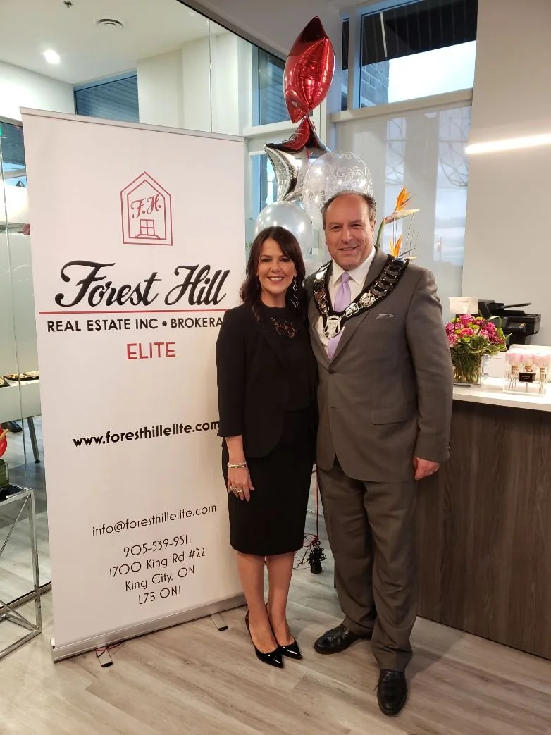 	
Mayor Steve Pellegrini is wearing a suit and tie, with a chain around his neck, and the woman is dressed in a black outfit. They are standing next to a sign for Forest Hill Real Estate Elite, with pink and white flowers on a counter nearby.