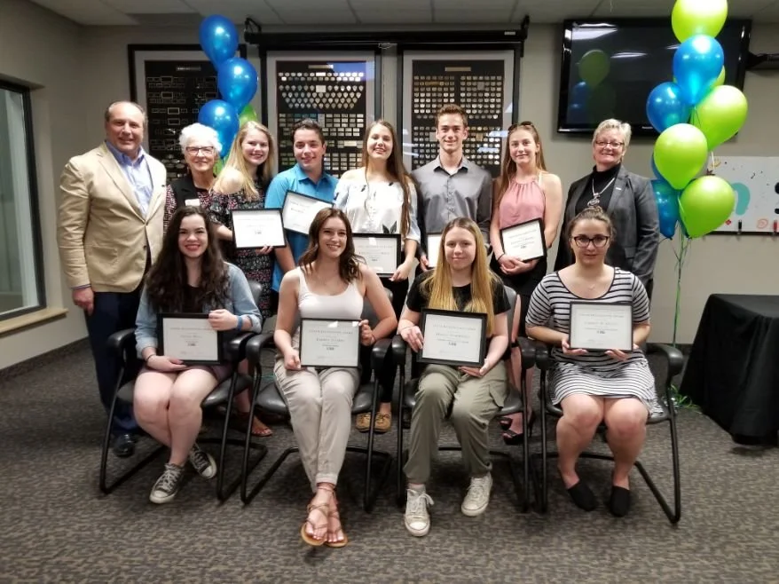 Group of young women and older adults at an award ceremony, holding certificates, with colorful balloons in the background.
