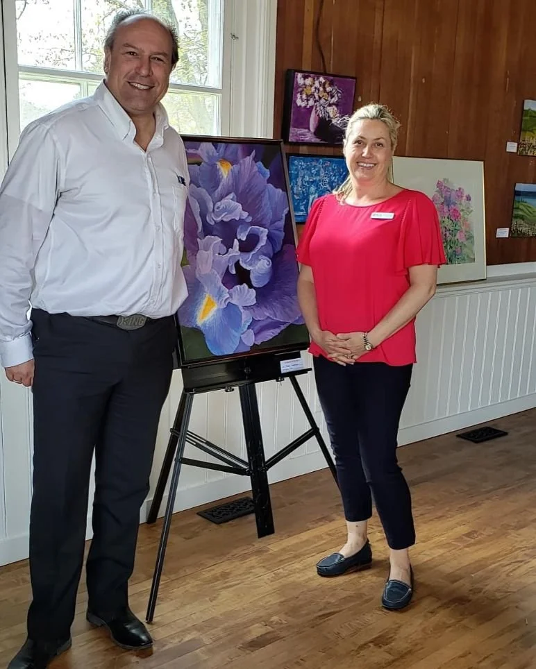 A man and a woman standing next to each other indoors at an art gallery, smiling, with a colorful purple and yellow iris painting on display on an easel between them. Additional paintings are visible on the wall behind them.