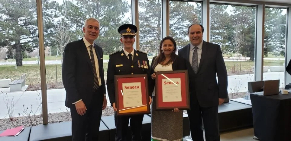 Four people standing together inside a room during a ceremony, with two of them holding framed certificates. One of the individuals is in a military uniform, and the others are dressed in formal attire. Large windows in the background reveal trees an