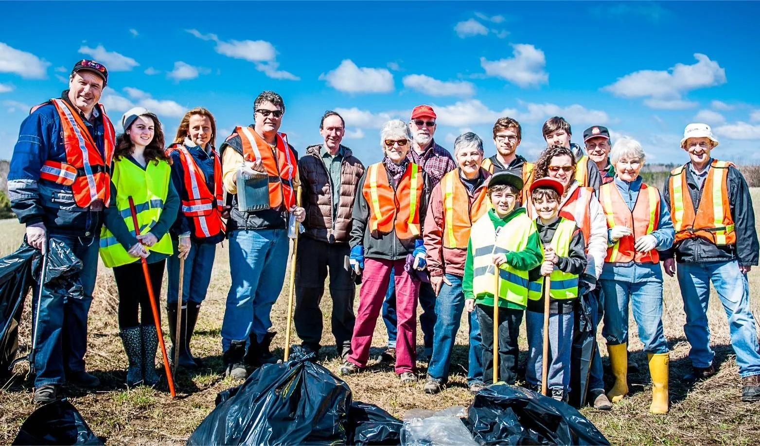 Mayor Steve Pellegrini participating in a community cleanup outdoors. They are wearing safety vests, holding trash pickers, and standing on a field with trash bags on the ground. The bright blue sky and clouds are in the background.