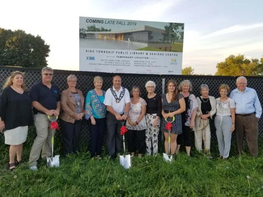 	
Mayor Steve Pellegrini standing in front of a fence with a sign announcing the coming of King Township Public Library & Seniors Centre, with trees in the background.