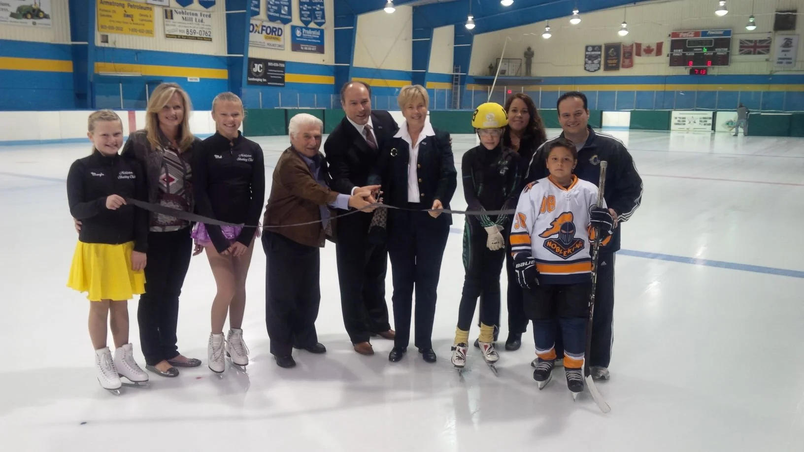Group of people at an ice skating rink during a ribbon-cutting ceremony, including children in skating gear and adults in formal and casual attire.