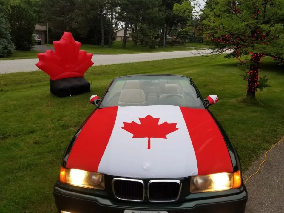 A black convertible car decorated with the Canadian flag paint and a large Canadian maple leaf inflatable on the lawn.