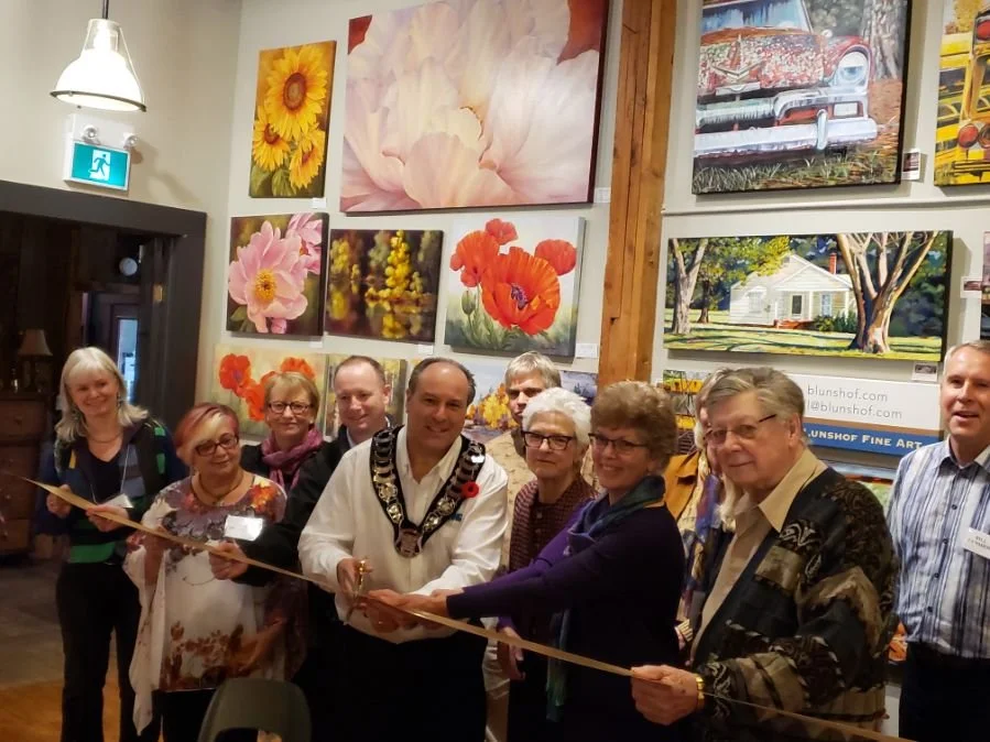 A group of people at an art gallery opening, standing in front of paintings of flowers and a house, participating in a ribbon-cutting ceremony.